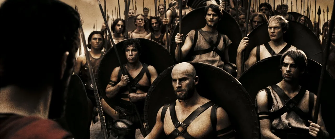 300 (2007) movie still: medium — A group of ancient warriors stands in formation holding spears and circular shields. The men have muscular builds…