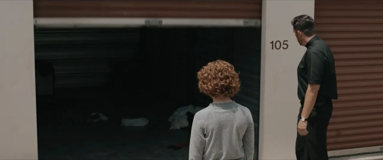 Possessions (2024) movie still: fullbody — A row of metal storage units with a partially open door numbered 105. A child and an adult man stand…
