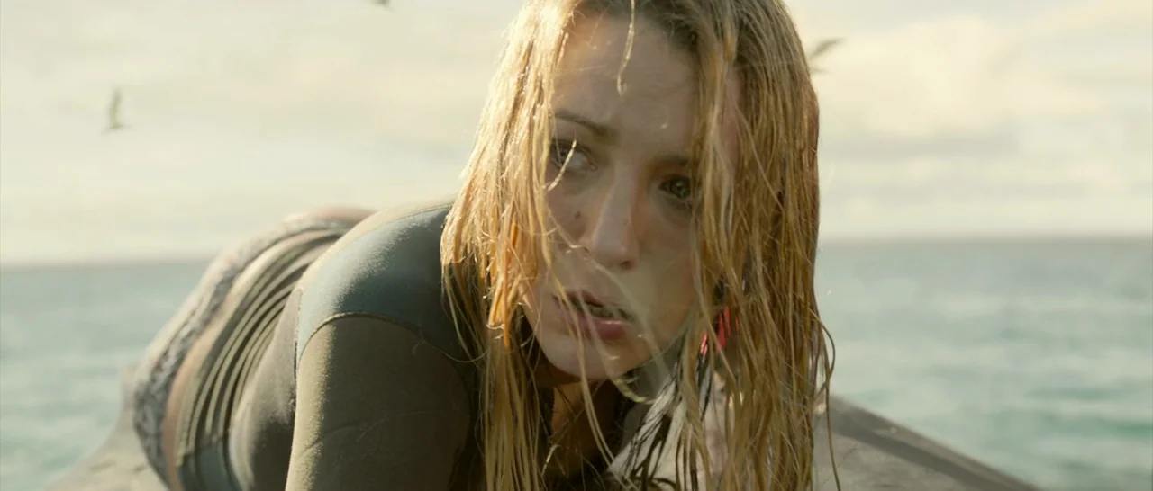 The Shallows (2016) movie still: closeup — A woman lies on a surfboard in the middle of a vast, bright ocean. She is looking back with a focused…