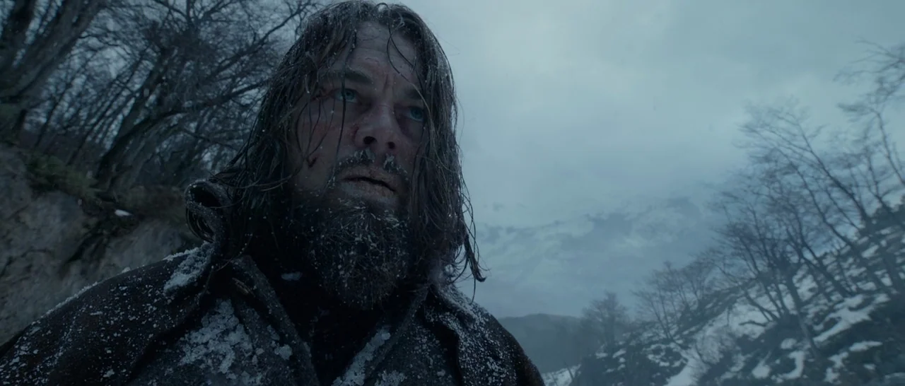 A man in a heavy coat stands amidst a snowy forest with bare trees. He stares forward while snow flakes collect on his hair and beard.