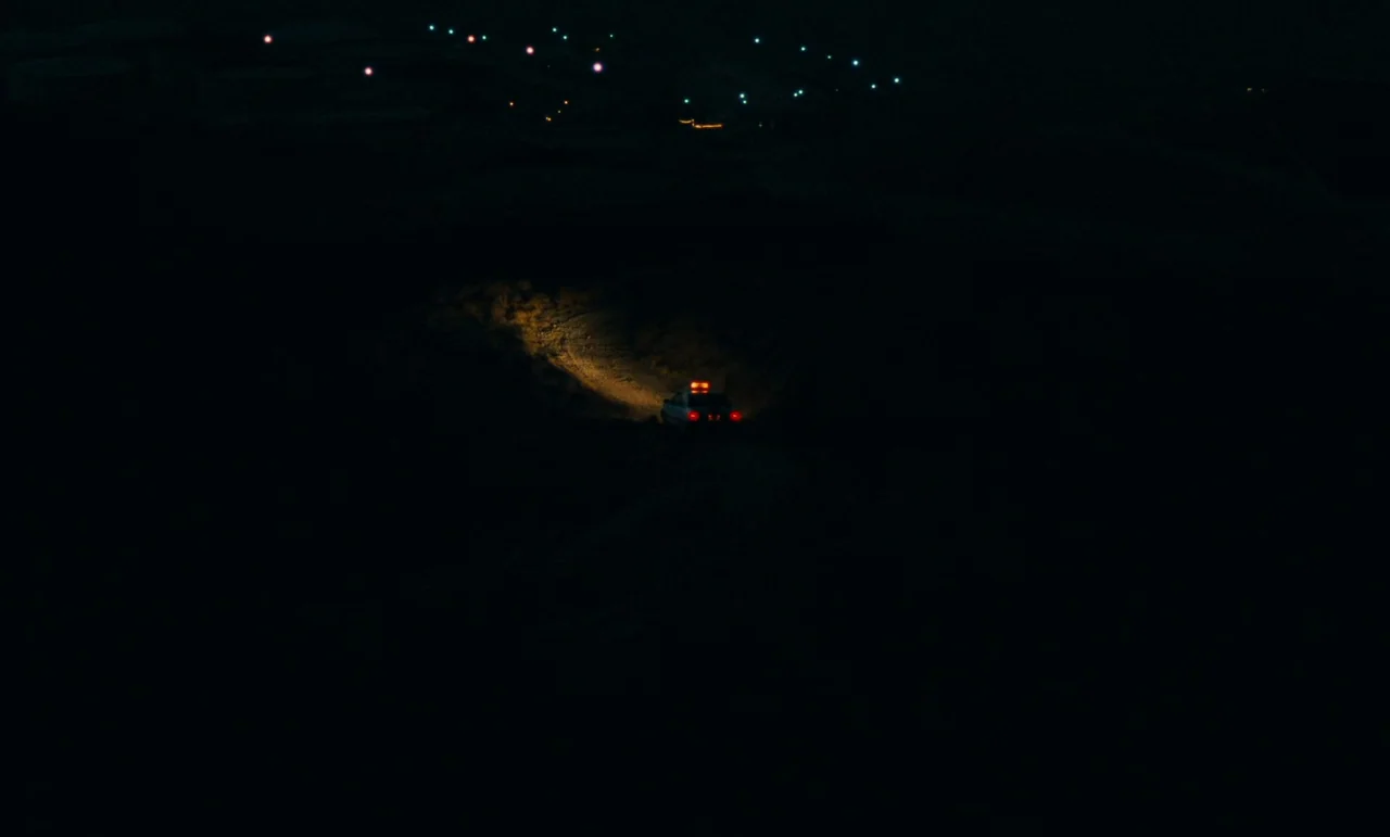 Taste of Cherry (1997) movie still: establishing — A vast dark desert landscape at night stretches under a faint distant city horizon. A single car…