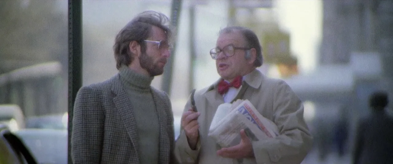 House By The Cemetery (1981) movie still: medium — A city street corner shows two men standing in conversation outdoors. One man gestures with a…
