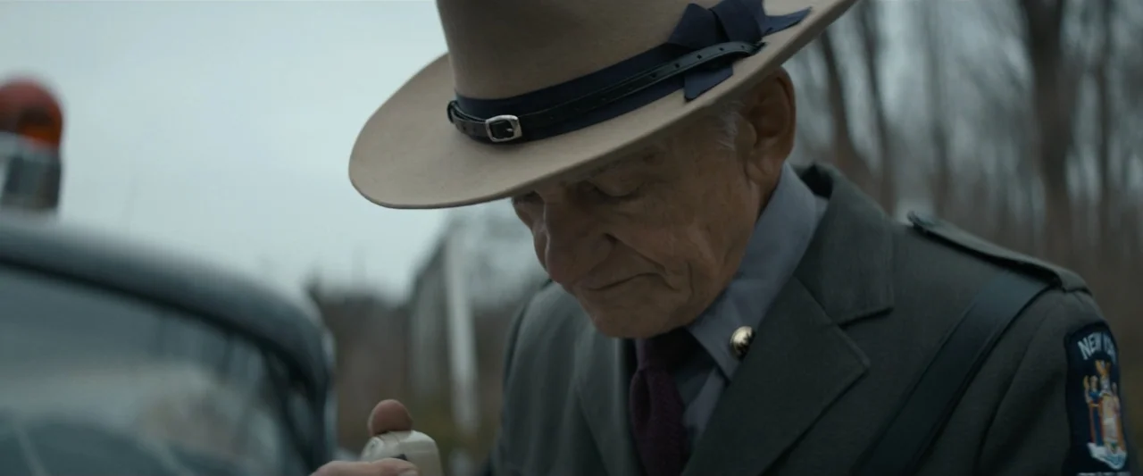 The Alto Knights (2025) movie still: closeup — An older state trooper in uniform stands outside in a cold overcast environment. He holds a police…