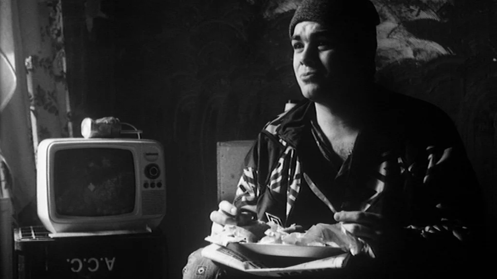 TwentyFourSeven (1997) movie still: medium — A dimly lit room contains a person seated beside an old television set. The person holds a plate of…