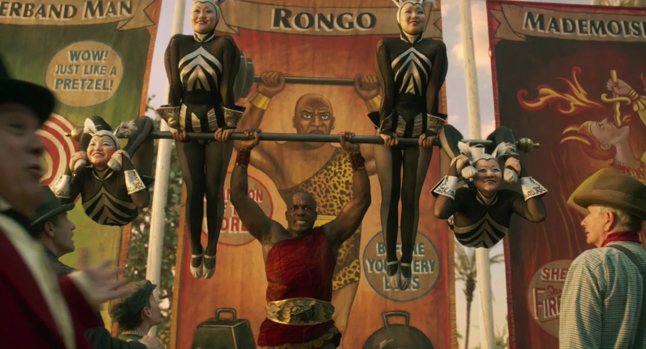 Dumbo (2019) movie still: medium — An outdoor circus performance features a strongman lifting a bar with four suspended acrobats. Large illustrated…