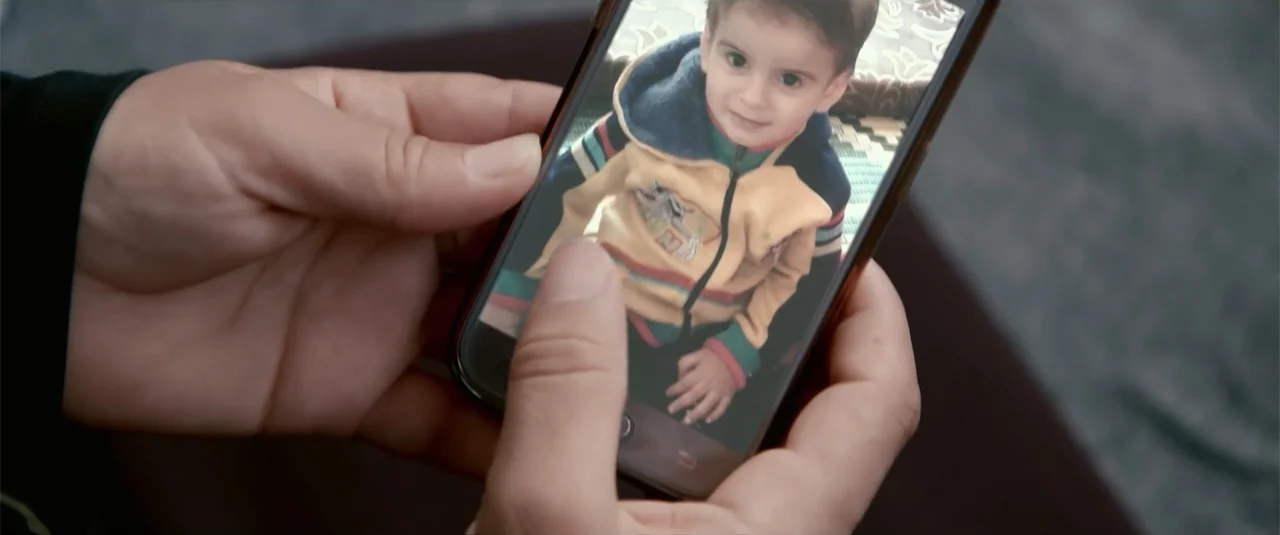Simple as Water (2021) movie still: closeup — A close-up view shows a person holding a smartphone showing a photo of a child. The child is pictured…