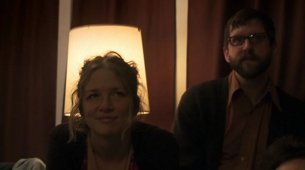 Your Sister’s Sister (2011) movie still: medium — A woman and a man sit in a darkened room with brown curtains behind them. A lit floor lamp between…