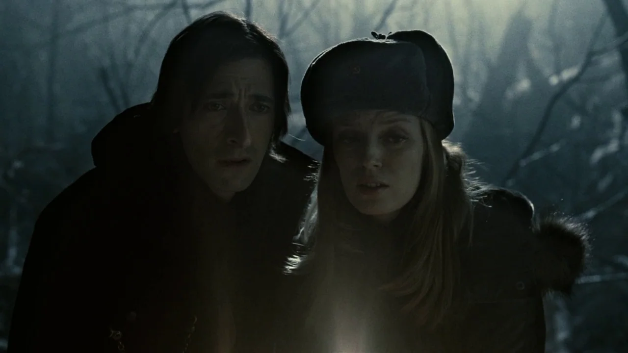 Splice (2009) movie still: medium — A dark, dense forest area at night with trees visible in the soft background. A man and a woman look down at an…