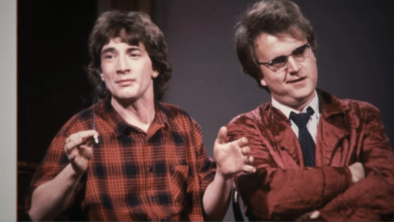 John Candy: I Like Me (2025) movie still: medium — A television studio set with a plain backdrop provides a minimalist interview environment. Two…