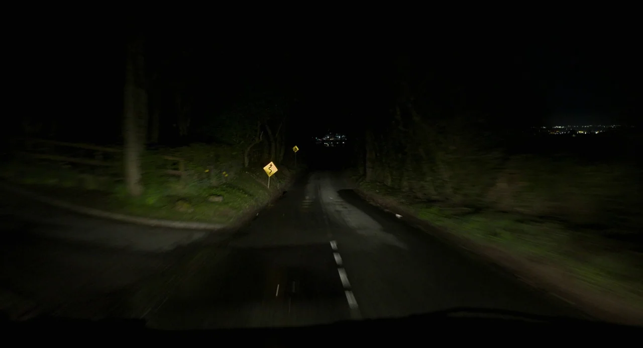 All You Need Is Death (2023) movie still: establishing — A dark, narrow country road at night is illuminated by approaching headlights. Two yellow…