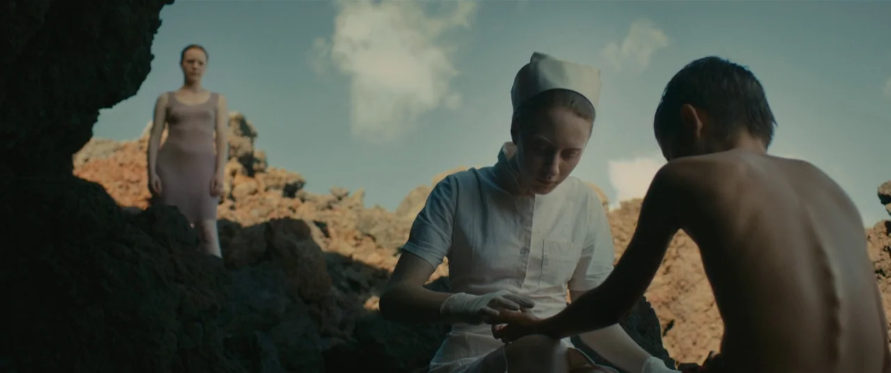 Evolution (2015) movie still: medium — A desolate landscape composed of dark, jagged volcanic rock under a bright sky. A nurse tends to a shirtless…