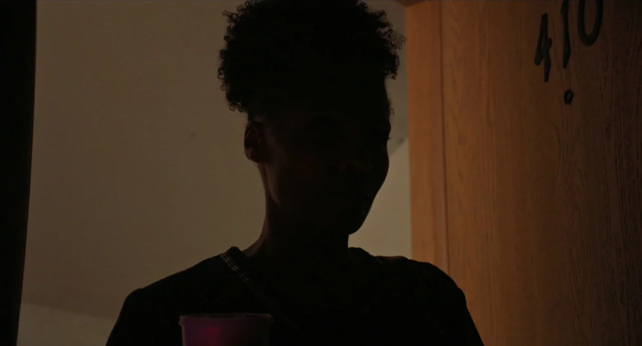Relaxer (2018) movie still: medium — A dimly lit hotel doorway features the number 410 on a wooden door. A person stands in the dark opening while…