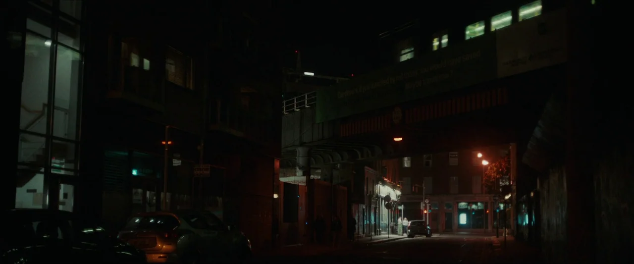 Fréwaka (2024) movie still: establishing — A dark city street passes under a massive concrete railway bridge at night. Several parked cars line the…