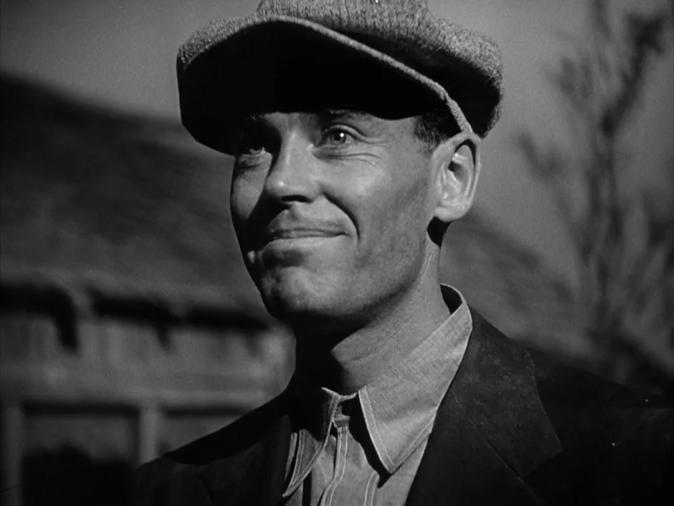 The Grapes of Wrath (1940) movie still: closeup — An outdoor field environment features a man posing against a blurred building structure. He wears…