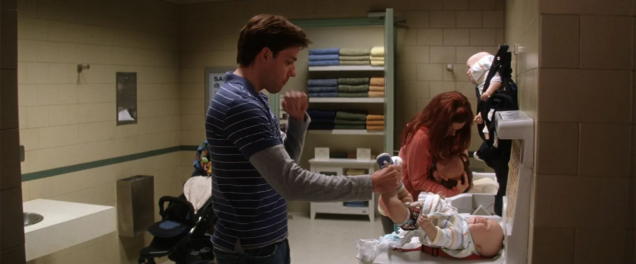 License to Wed (2007) movie still: medium — A utilitarian public restroom with tiled walls features a shelving unit stacked with towels. A man and a…