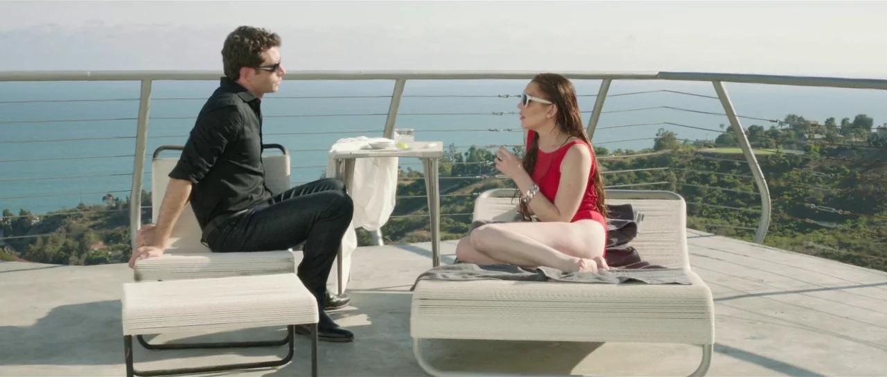 The Canyons (2013) movie still: wide — A modern cliffside terrace overlooks a distant ocean with a simple metal wire railing. A man and a woman sit…