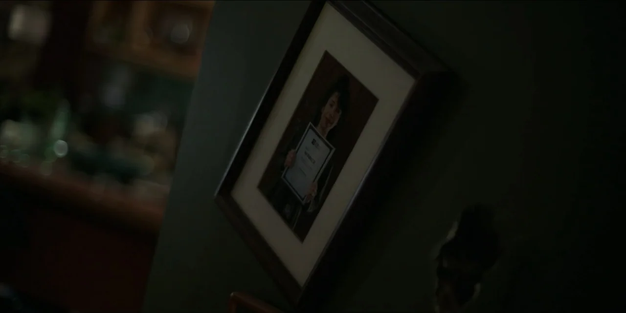 Bring Her Back (2025) movie still: closeup — A dark interior wall features a crookedly mounted framed photograph. The image inside the frame…