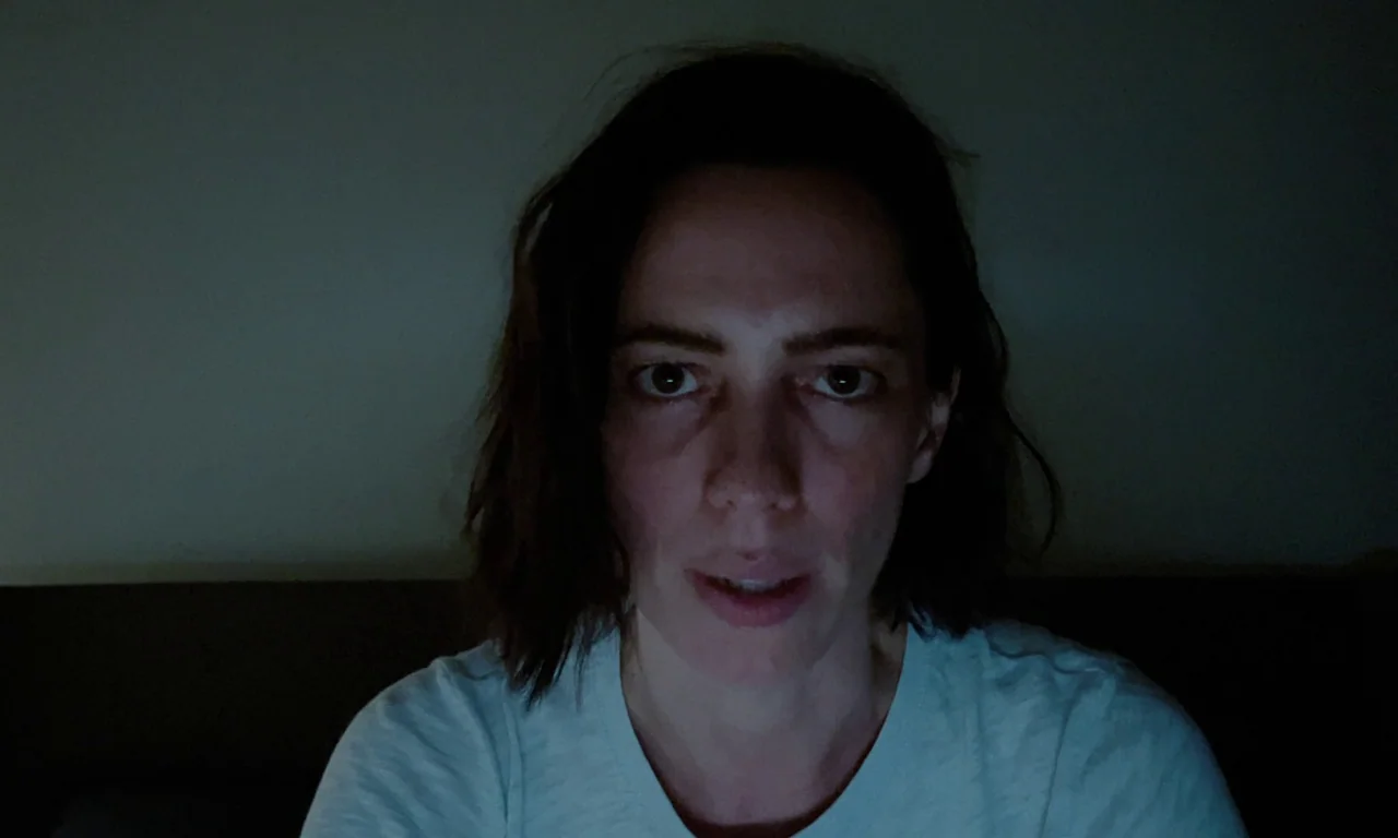 Resurrection (2022) movie still: closeup — A woman is centered in a dark interior space against a featureless wall. She faces forward and appears to…