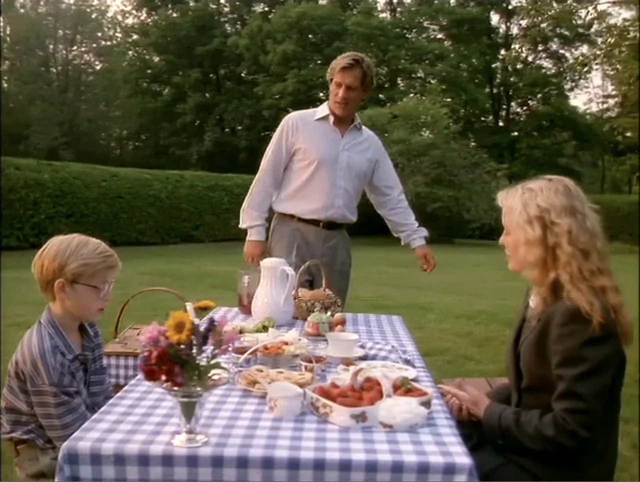 At the Midnight Hour (1995) movie still: fullbody — A picnic table covered in a blue and white checkered cloth sits on a lawn. A man stands behind…