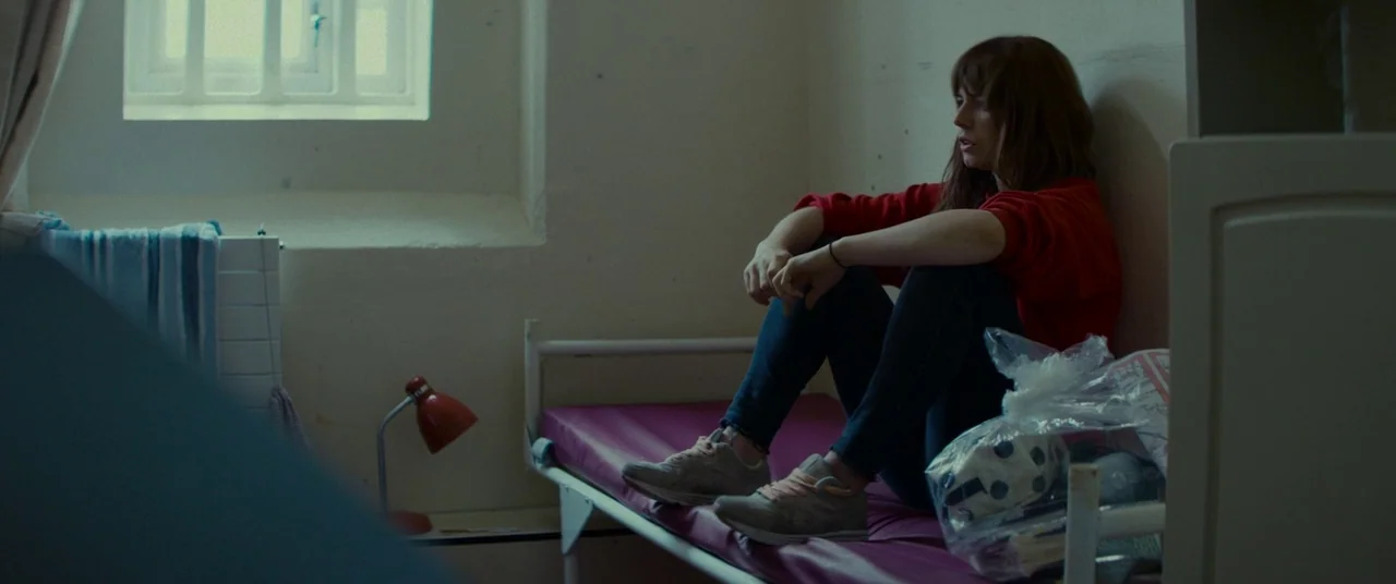 Wild Rose (2018) movie still: medium — A spartan room features beige walls, a small high window with bars, and a cot. A young woman sits on the bed…