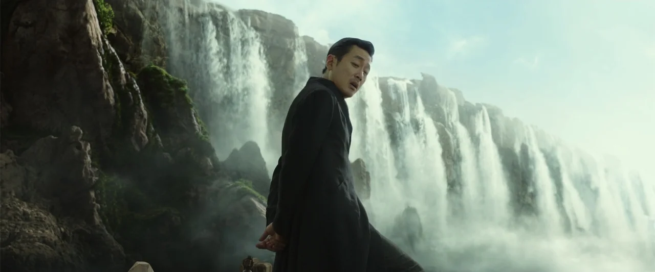 Along with the Gods: The Last 49 Days (2018) movie still: medium — A man stands before a large, misty waterfall crashing against rocky cliffs. The…