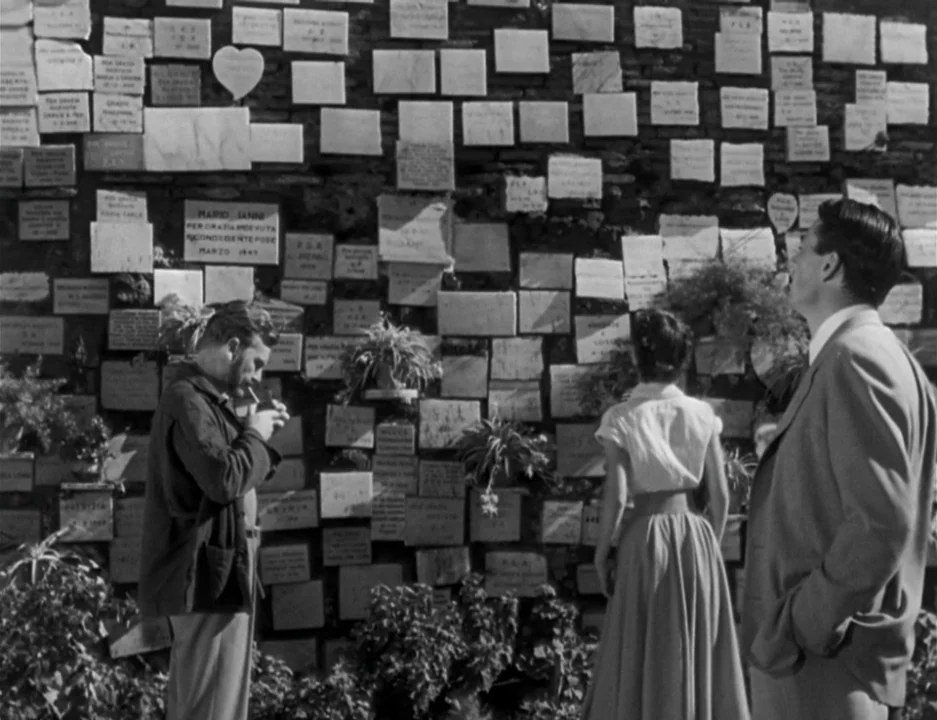 Roman Holiday (1953) movie still: fullbody — A stone brick wall is densely covered with rectangular white memorial plaques and small plants. Three…