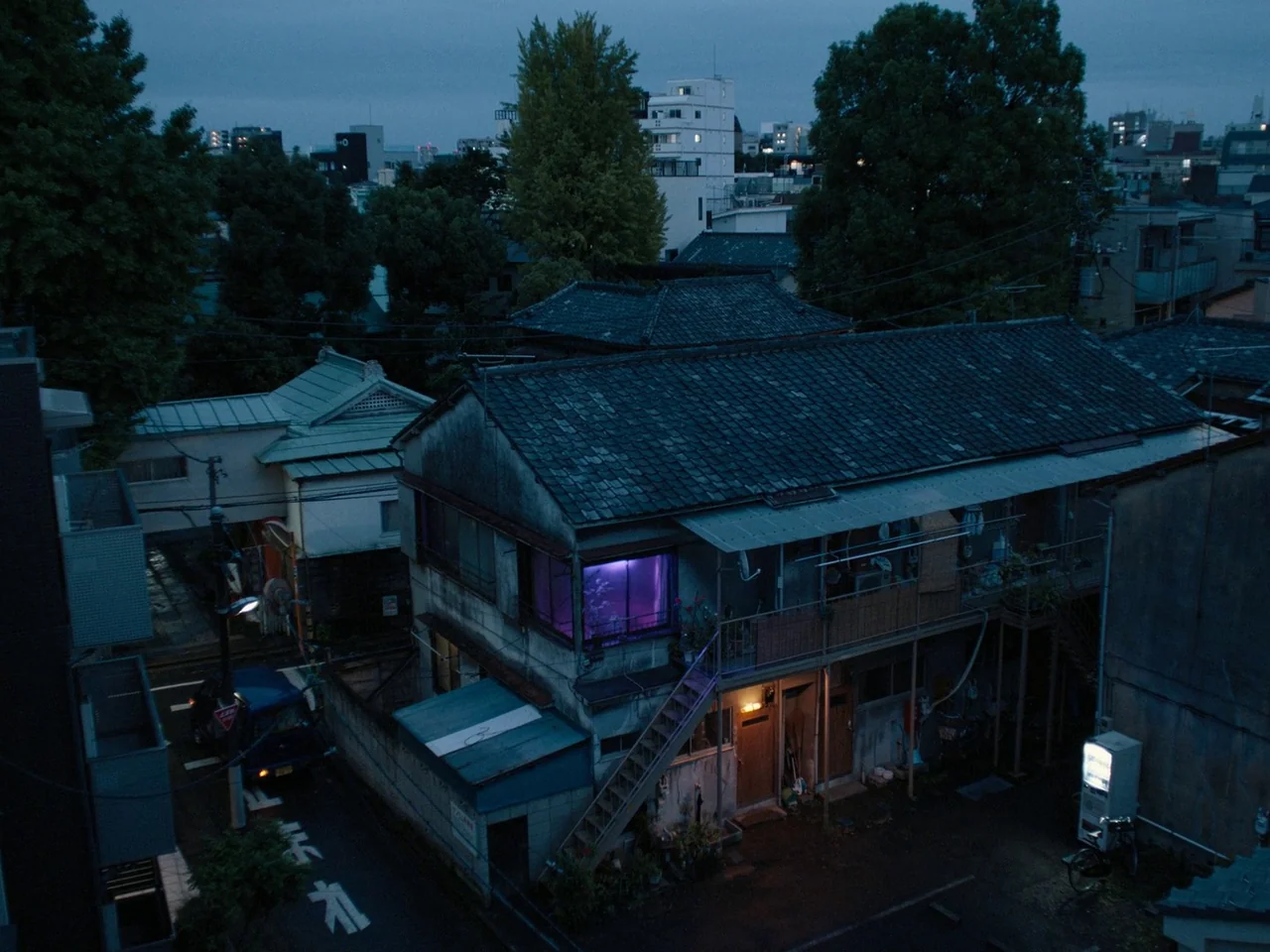 Perfect Days (2023) movie still: establishing — An aging two-story apartment building sits on a quiet street in a Japanese neighborhood. The…