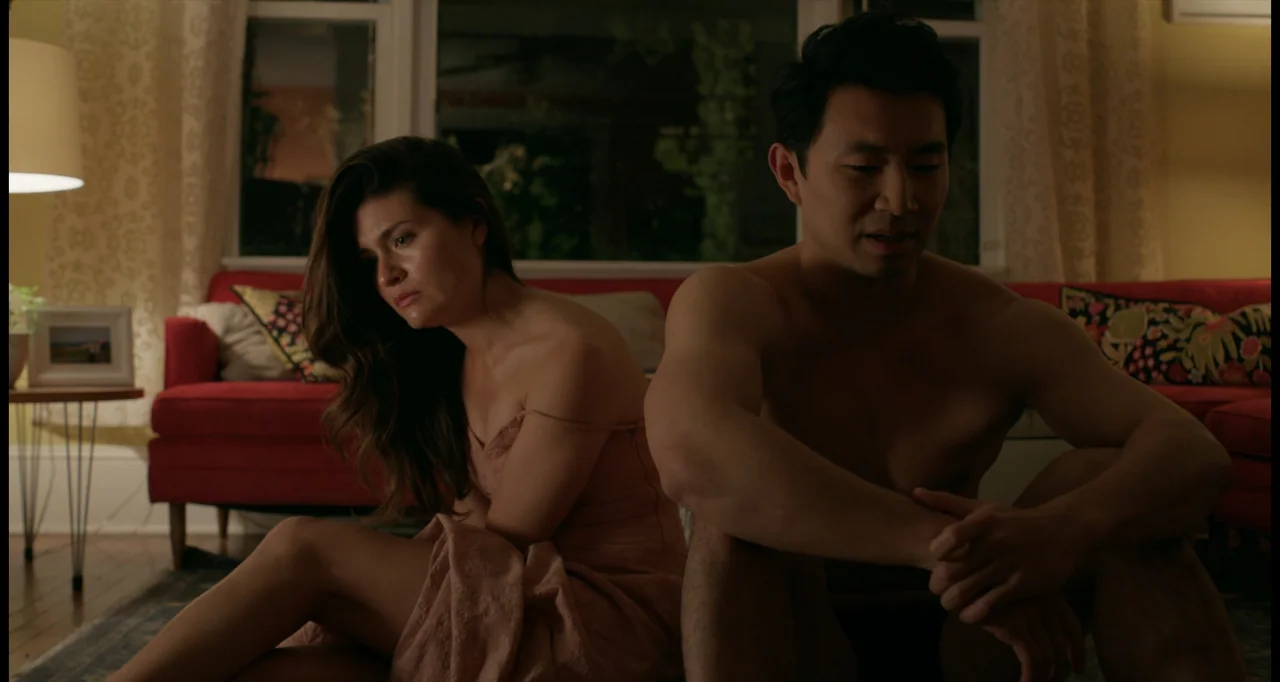 One True Loves (2023) movie still: medium — A living room interior featuring a red couch and a large dark window at night. A man and a woman sit on…