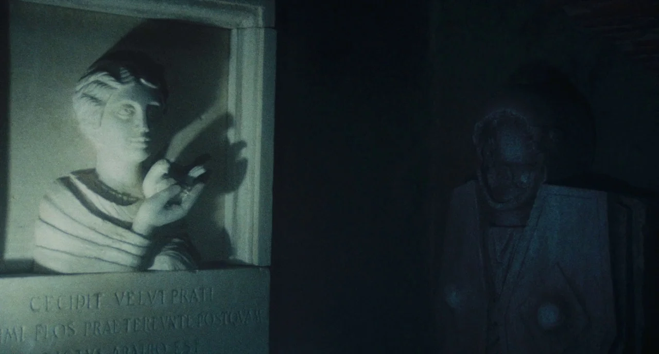 Roma (1972) movie still: medium — A dark stone crypt contains a carved marble wall plaque featuring a female bust. The bust is illuminated by a…