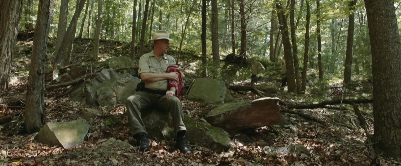 Darkness Waits (2020) movie still: medium — A dense forest clearing features uneven rocky ground covered in fallen autumn leaves. A park ranger in…