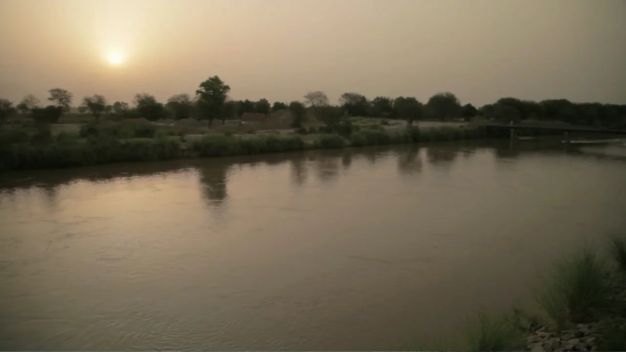 A Girl in the River: The Price of Forgiveness (2015) movie still: establishing — A wide river flows through an open rural landscape during a hazy…