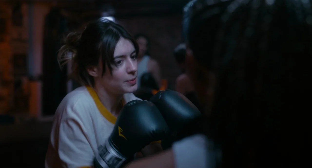 Fresh (2022) movie still: medium — A dimly lit training space with dark, out-of-focus background textures. A woman in a white t-shirt and boxing…