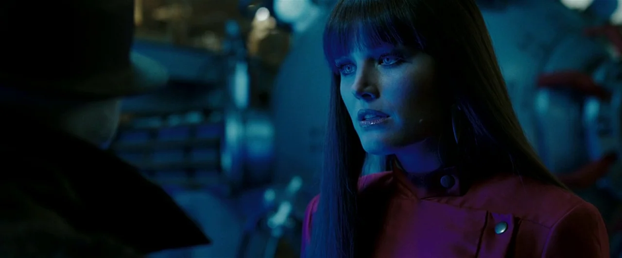 Watchmen (2009) movie still: closeup — A dark, blue-lit industrial space with mechanical equipment visible in the blurred background. A woman with…