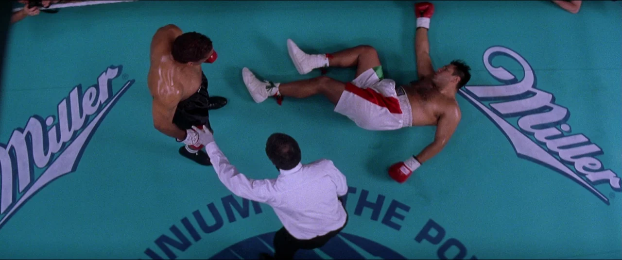 Snake Eyes (1998) movie still: wide — A top-down view of a turquoise canvas with Miller logos in a large arena. A referee hovers over a fallen boxer…