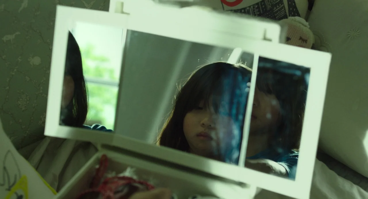 The Mimic (2017) movie still: closeup — A child sits in a room with light green wallpaper while looking into a vanity mirror. The reflection shows…