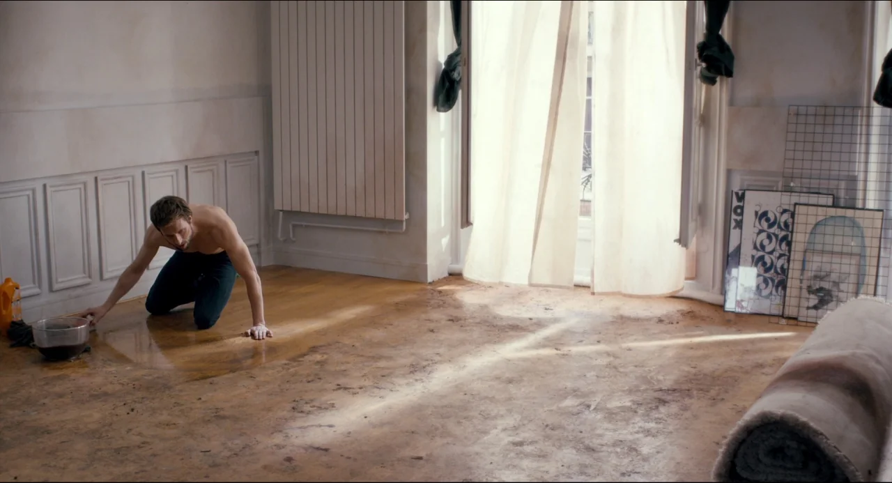 The Night Eats the World (2018) movie still: wide — A bright room has light hardwood flooring covered in dirt and dark liquid spills. A shirtless…