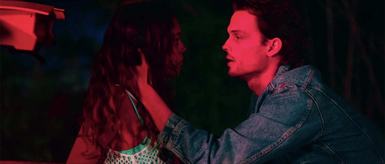 68 Kill (2017) movie still: medium — An outdoor location at night with dark surroundings and heavy red-tinted lighting. A young man reaches out to…