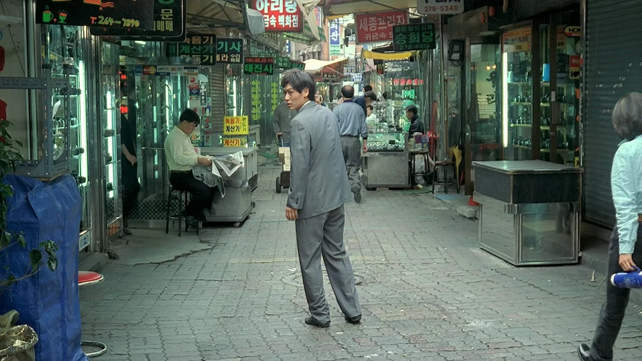 Peppermint Candy (1999) movie still: fullbody — A dense urban market alleyway features numerous small shop fronts adorned with Korean signage. A man…