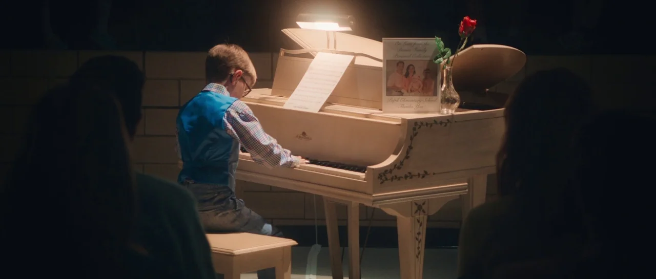 Greener Grass (2019) movie still: medium — A dimly lit indoor auditorium featuring a white grand piano as the central focus. A young boy in a vest…