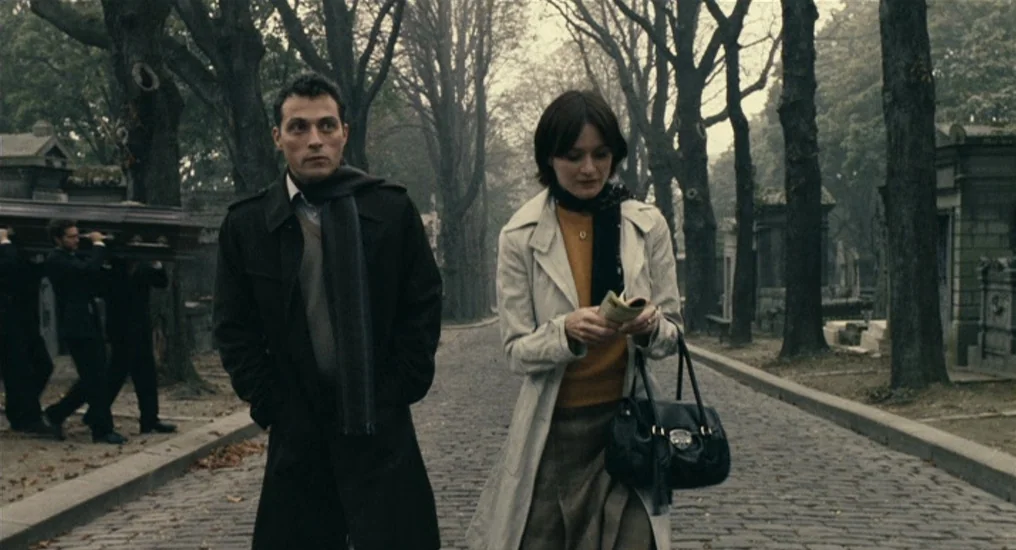 Paris, Je T’Aime (2006) movie still: fullbody — A historic cemetery with rows of mature trees flanking a narrow cobblestone path. A man and woman…