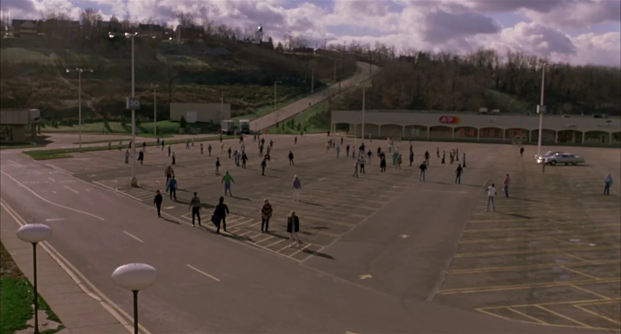 Dawn of the Dead (1978) movie still: establishing — A vast, paved parking lot stretches toward a low-rise building under a cloudy sky. Many people…