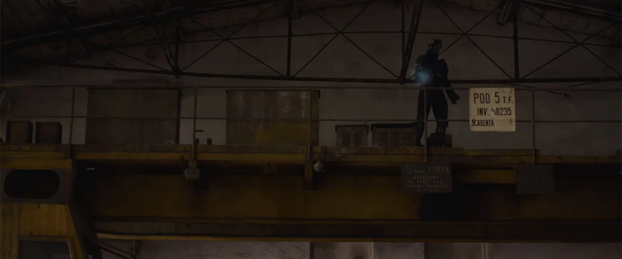 The Condemned 2 (2015) movie still: wide — An industrial catwalk overlooks a darkened space with heavy metal machinery and signage. A solitary…