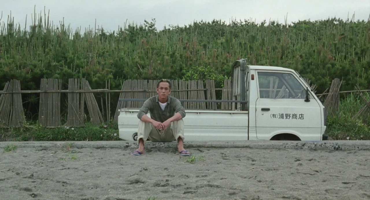 A Scene at the Sea (1991) movie still: fullbody — A man sits on a low concrete curb near a white utility truck. Tall grasses and a bamboo fence form…