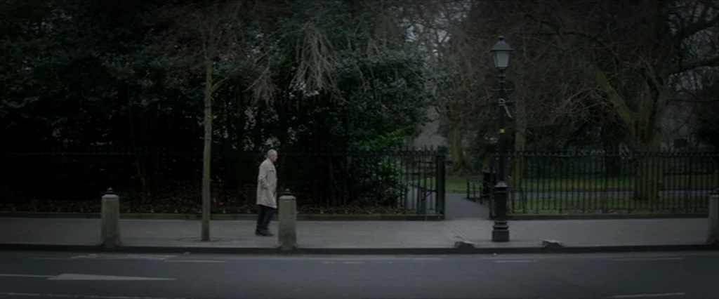 Haywire (2011) movie still: wide — A man walks past a black iron fence bordering a park in the city. The sidewalk is lined with trees and a single…