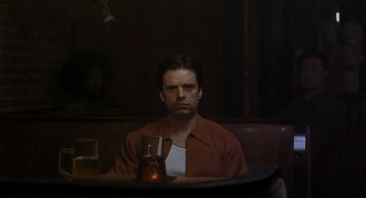 A Different Man (2024) movie still: medium — A dimly lit restaurant booth with a brick wall and wooden paneling in the background. A man sits alone…