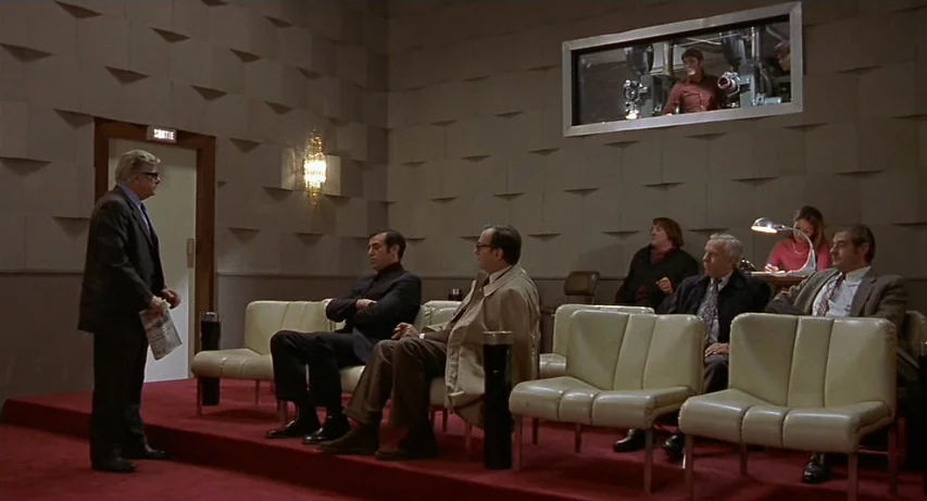 CQ (2001) movie still: wide — A modern screening room with geometric wall paneling and rows of beige plastic chairs. A group of men sits in the room…