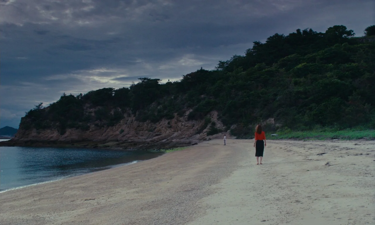 Sidonie in Japan (2023) movie still: wide — A secluded coastal beach stretches beneath a heavy, dark overcast sky. A woman walks barefoot along the…
