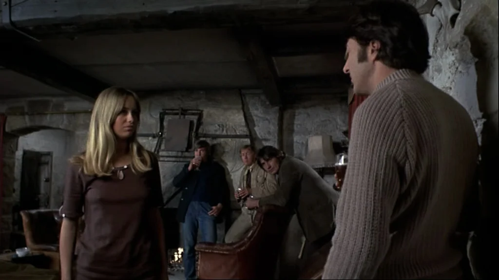 Straw Dogs (1971) movie still: medium — A rustic pub interior features rough stone walls, heavy timber beams, and dim lighting. Several people…