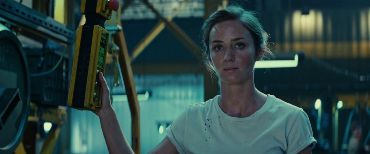 Edge of Tomorrow (2014) movie still: medium — A dimly lit industrial environment features metallic structures and hanging equipment. A woman stands…