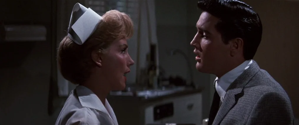 It Happened At The World’s Fair (1963) movie still: medium — A dimly lit room with medical station cabinetry visible in the background shadow. A…